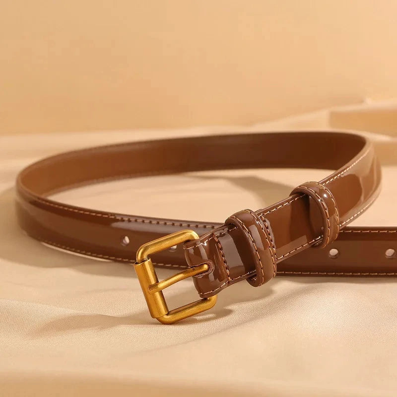 cinturón marrón de charol con hebilla dorada parcialmente enrollado sobre superficie beige